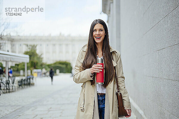 Lachende Frau mit isoliertem Getränkebehälter  die in der Nähe einer Mauer in der Stadt steht