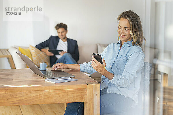 Lächelnde Frau mit Laptop auf dem Tisch  die ihr Smartphone hält  während ihr Freund zu Hause im Büro sitzt