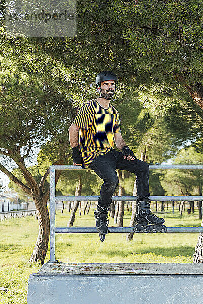 Mittlerer erwachsener Mann mit Sporthelm und Rollschuhen auf einem Geländer im Park sitzend