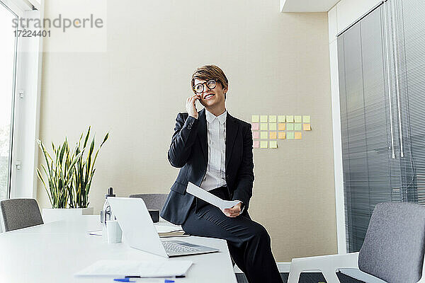 Lächelnde weibliche Fachkraft  die wegschaut  während sie am Schreibtisch mit einem Mobiltelefon spricht