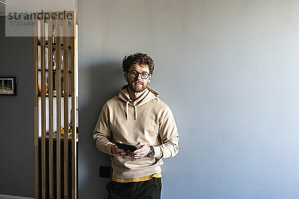 Mann mit Brille hält digitales Tablet und lehnt sich zu Hause an die Wand