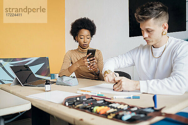 Junge Frau benutzt ihr Smartphone  während ein Mann am Tisch im Studio arbeitet