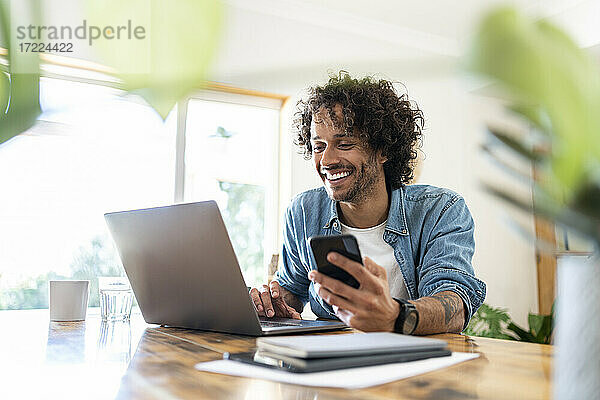 Lächelnder männlicher Berufstätiger  der ein Smartphone hält  während er einen Laptop auf einem Tisch zu Hause benutzt
