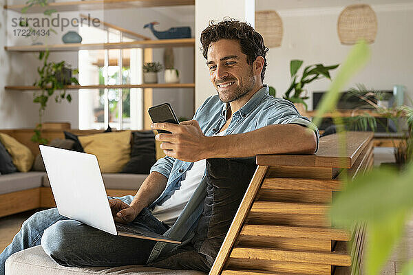 Lächelnder männlicher Freiberufler mit Laptop und Smartphone auf der Couch im Wohnzimmer