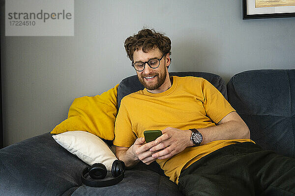 Lächelnder Mann  der ein Mobiltelefon benutzt  während er zu Hause auf dem Sofa sitzt