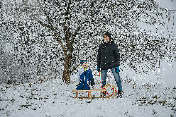 Vater und Sohn stehen im Winter mit Schlitten vor einem Baum