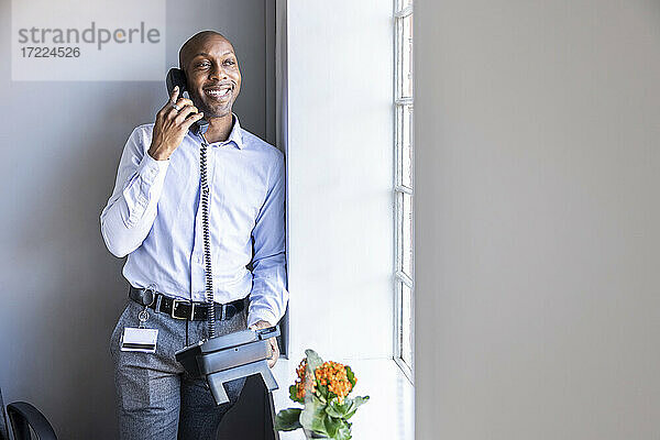 Lächelnder männlicher Unternehmer  der am Festnetztelefon spricht und sich im Büro an die Wand lehnt