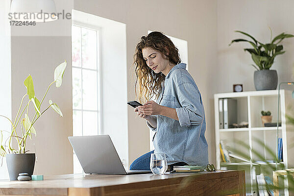 Geschäftsfrau mit Mobiltelefon  die eine Textnachricht schreibt  während sie auf dem Schreibtisch im Heimbüro sitzt