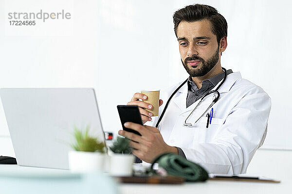 Gutaussehender männlicher Angestellter im Gesundheitswesen  der Kaffee trinkt und ein Mobiltelefon benutzt