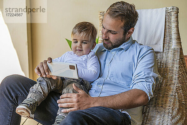 Vater teilt Mobiltelefon mit süßem Sohn  der zu Hause auf dem Schoß sitzt