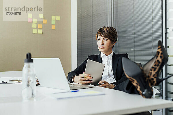 Nachdenkliche Geschäftsfrau mit hochgelegten Füßen und digitalem Tablet am Schreibtisch im Büro