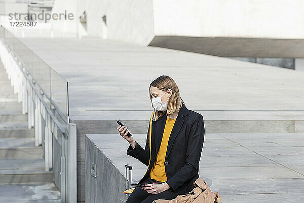 Unternehmerin mit Gesichtsmaske  die ein Mobiltelefon benutzt  während sie an einer Treppe sitzt