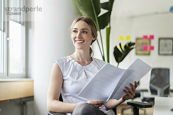 Nachdenkliche Geschäftsfrau mit Dokumenten  die lächelnd in ein Büro blickt