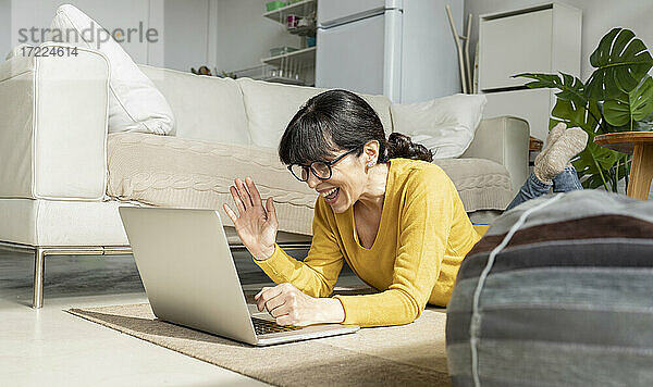 Erstaunte Frau gestikuliert während eines Videogesprächs über einen Laptop  während sie zu Hause auf dem Boden liegt