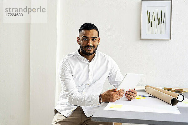 Männlicher Unternehmer mit digitalem Tablet sitzt am Schreibtisch an der Wand im Büro