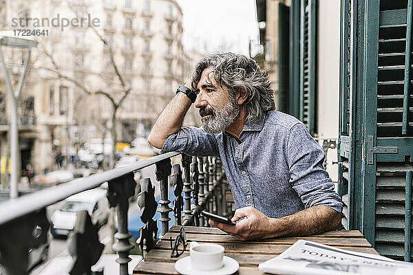 Nachdenklicher älterer Mann mit Smartphone sitzt am Tisch auf dem Balkon eines Hauses