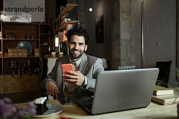 Glücklicher Geschäftsmann mit Laptop und Mobiltelefon in einem Café sitzend