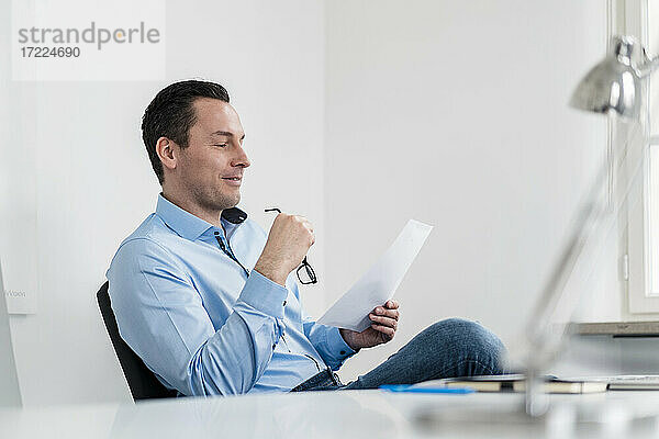 Männlicher Unternehmer hält eine Brille  während er am Arbeitsplatz ein Dokument liest