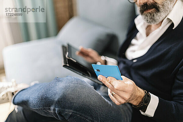 Ein älterer Mann hält eine Kreditkarte in der Hand  während er ein digitales Tablet zu Hause benutzt