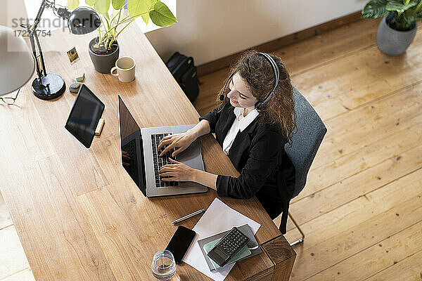 Weibliche Kundenbetreuerin mit Kopfhörern  die mit einem Kunden spricht  während sie im Büro zu Hause am Laptop arbeitet