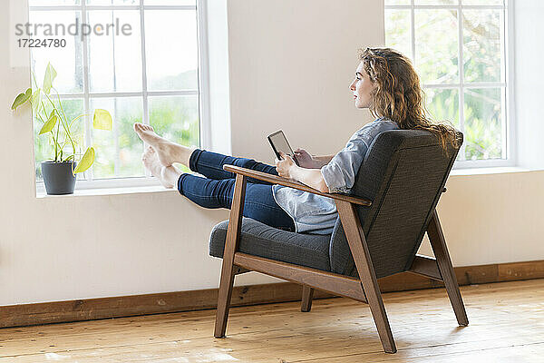 Junge Frau hält digitales Tablet  während sie zu Hause auf einem Sessel sitzt