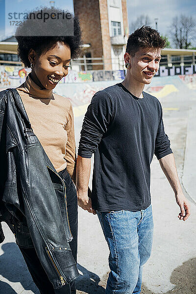 Lächelndes multiethnisches Paar  das in einem Skateboard-Park an einem sonnigen Tag spazieren geht und die Hände hält