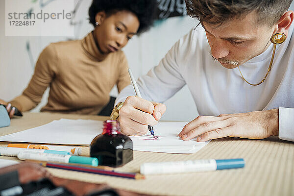 Hipster-Mann schreibt mit Füllfederhalter auf Papier  während er neben einer Frau im Atelier sitzt