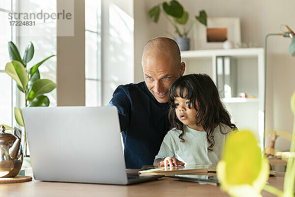 Vater arbeitet am Laptop  während er mit seinem Kleinkind im Home Office sitzt