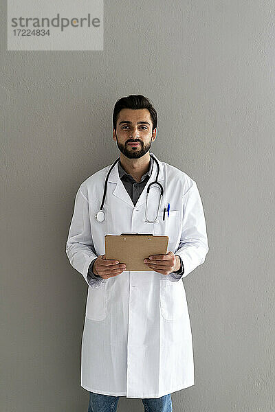 Männlicher Arzt mit Klemmbrett vor einer Wand stehend