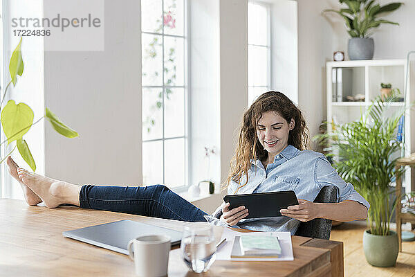 Lächelnde Unternehmerin  die auf ein digitales Tablet schaut  während sie mit hochgelegten Füßen am Schreibtisch sitzt