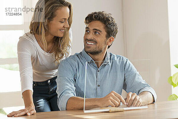 Glückliches Paar  das sich gegenseitig ansieht  während es mit einem transparenten Bildschirm zu Hause sitzt
