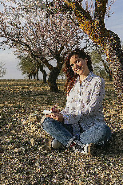 Lächelnde junge Frau sitzt mit Notizblock unter einem Mandelbaum