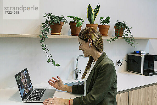 Fröhliche Geschäftsfrau mit In-Ear-Kopfhörern bei einem Videogespräch am Laptop im Büro