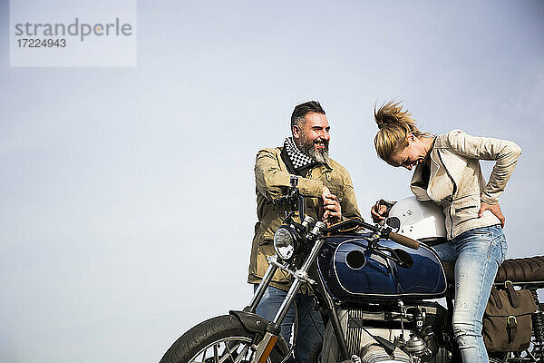 Lächelnder Mann im Gespräch mit fröhlicher Bikerin auf Motorrad sitzend am Himmel