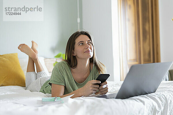 Nachdenkliche junge Frau mit Smartphone  die zu Hause auf dem Bett vor dem Laptop liegt