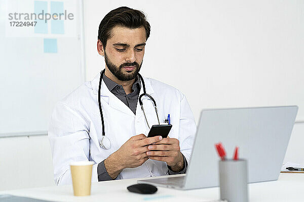Männlicher Arzt  der am Schreibtisch sitzend ein Mobiltelefon benutzt