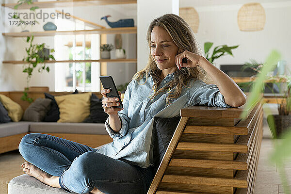 Blondhaarige Frau  die im Wohnzimmer sitzend eine SMS über ihr Smartphone verschickt