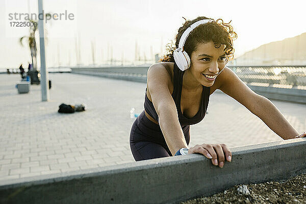 Lächelnde Sportlerin hört Musik über Kopfhörer  während sie auf einer Brücke trainiert