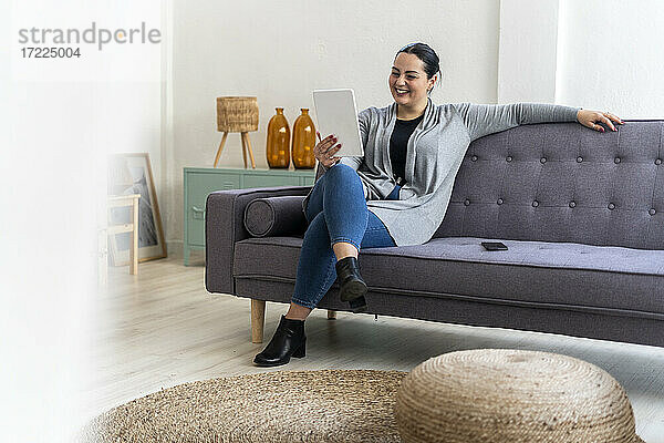 Lächelnde Frau  die ein digitales Tablet benutzt  während sie auf dem Sofa im Wohnzimmer sitzt