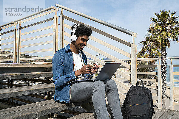 Glücklicher Mann gestikuliert während eines Videogesprächs am Laptop  während er an einem sonnigen Tag auf der Promenade sitzt