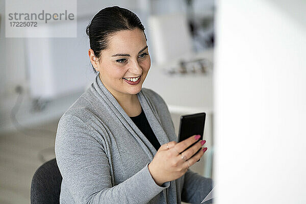 Lächelnde weibliche Fachkraft  die im Büro zu Hause ein Mobiltelefon benutzt
