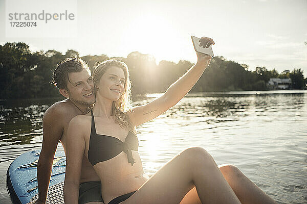 Glückliches Paar sitzt auf einem Paddelbrett auf einem See und macht Smartphone-Selfies
