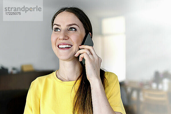 Glückliche Frau mit grauen Augen  die wegschaut  während sie zu Hause mit dem Handy telefoniert