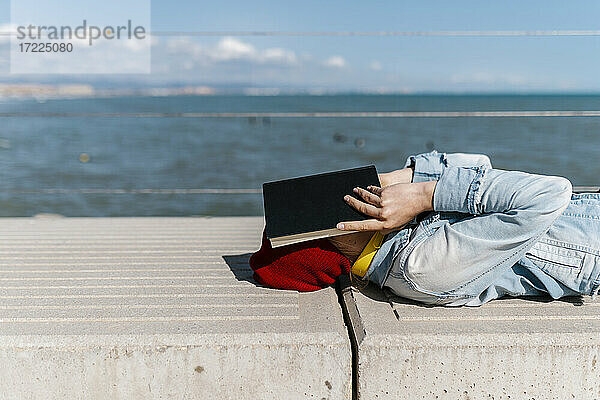 Junge Frau  die ihr Gesicht mit einem Tagebuch bedeckt  während sie sich an einem sonnigen Tag auf eine Stützmauer legt