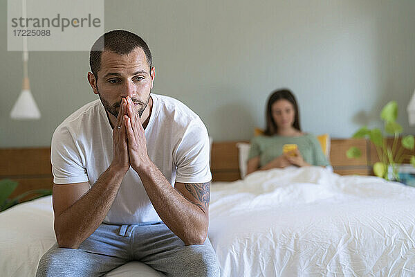 Besorgter junger Mann sitzt auf dem Bett  während seine Freundin im Hintergrund ihr Smartphone benutzt