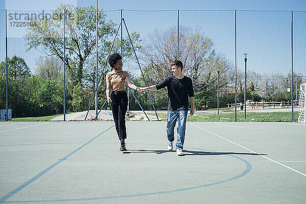 Glückliches multiethnisches Paar  das an einem sonnigen Tag auf einem Sportplatz spazieren geht und die Hände hält