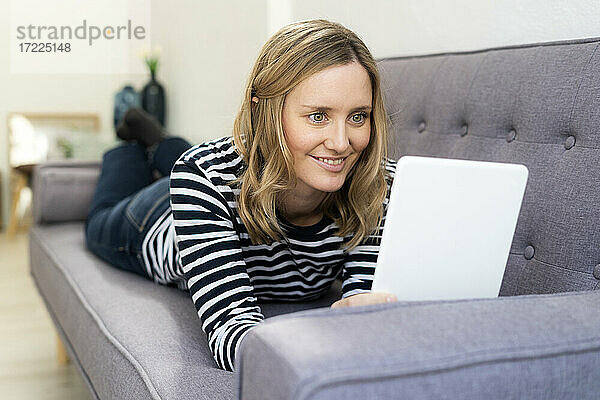 Lächelnde Frau  die ein digitales Tablet benutzt  während sie zu Hause auf dem Sofa liegt