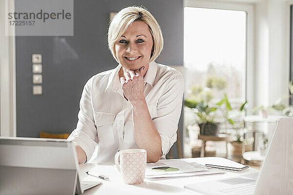 Lächelnde Unternehmerin mit digitalem Tablet und Laptop am Tisch im Heimbüro sitzend