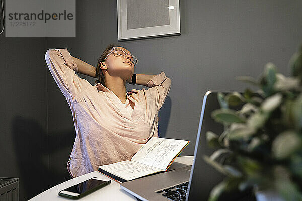 Müde Frau sitzt mit den Händen hinter dem Kopf am Laptop zu Hause