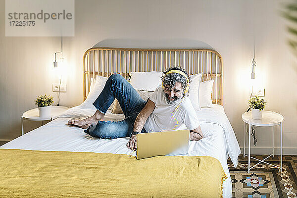 Entspannter älterer Mann mit Kopfhörern und Laptop auf dem Bett zu Hause